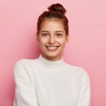 tender feminine woman with blue eyes smiles pleasantly has toothy smile wears white comfortable sweater looks directly camera isolated pink background