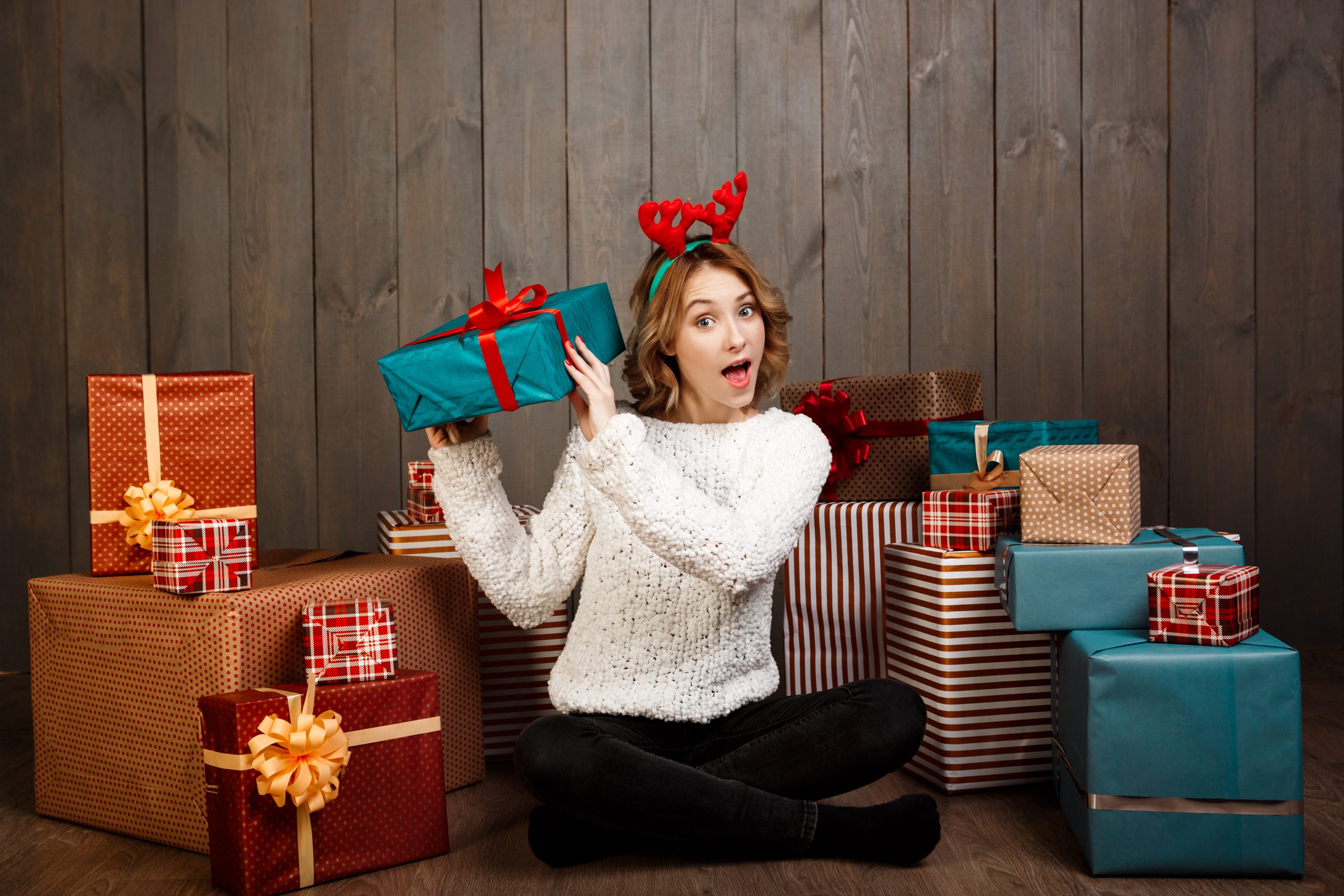 young beautiful girl sitting among christmas gifts wooden wall scaled